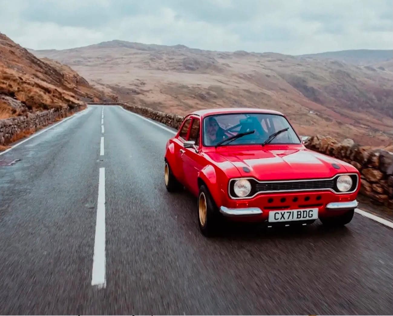 Red Ford Escort Mk1 rally build on a wet mountain pass, gold wheels, roll cage visible through the glass