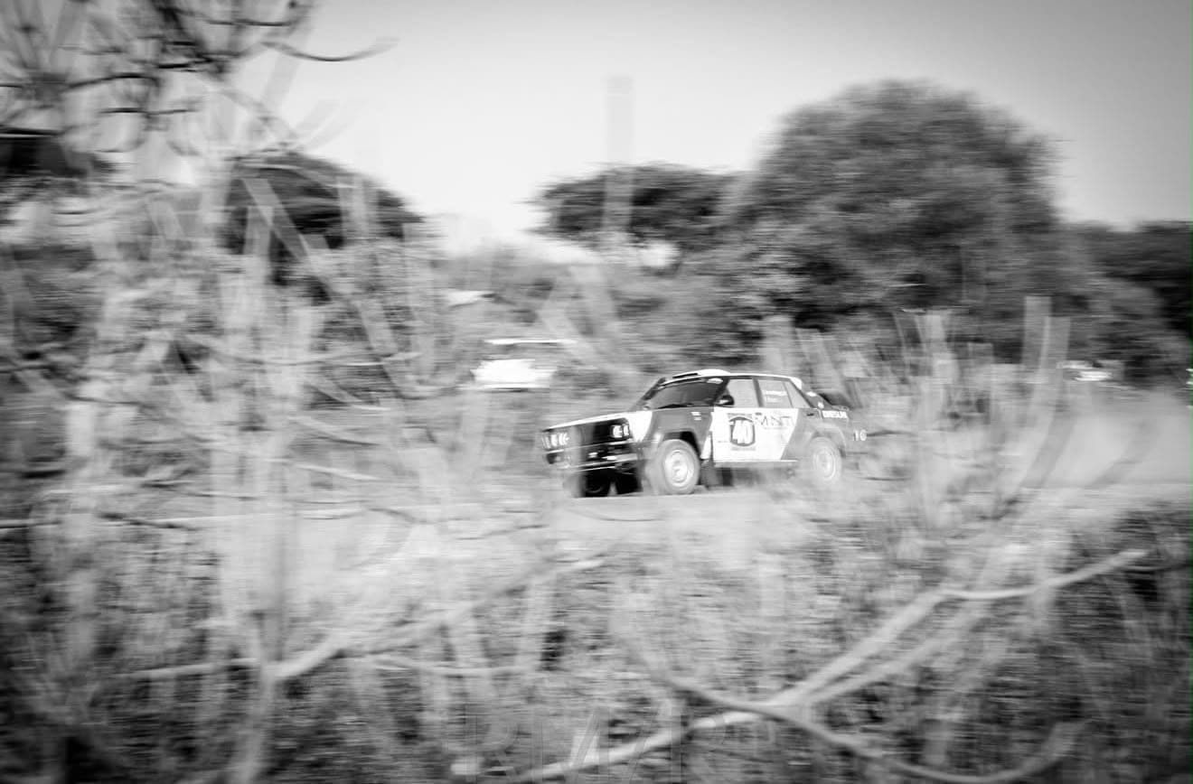 Archive black-and-white photograph: Minti-liveried classic rally saloon at speed on an East African stage, panned through thorn scrub
