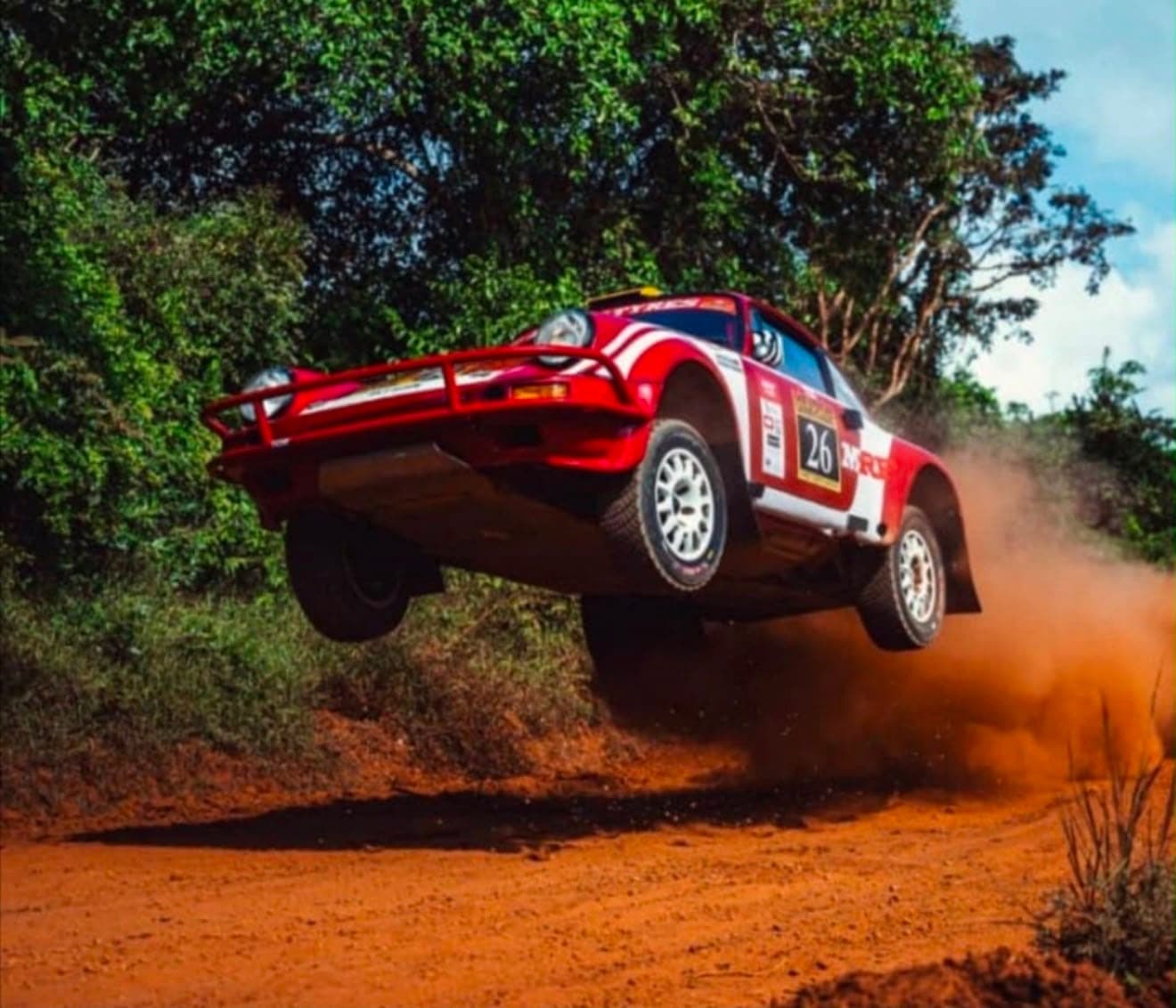 Safari rally car, competition number 26, airborne over a murram crest, red dust trailing, East African bush behind